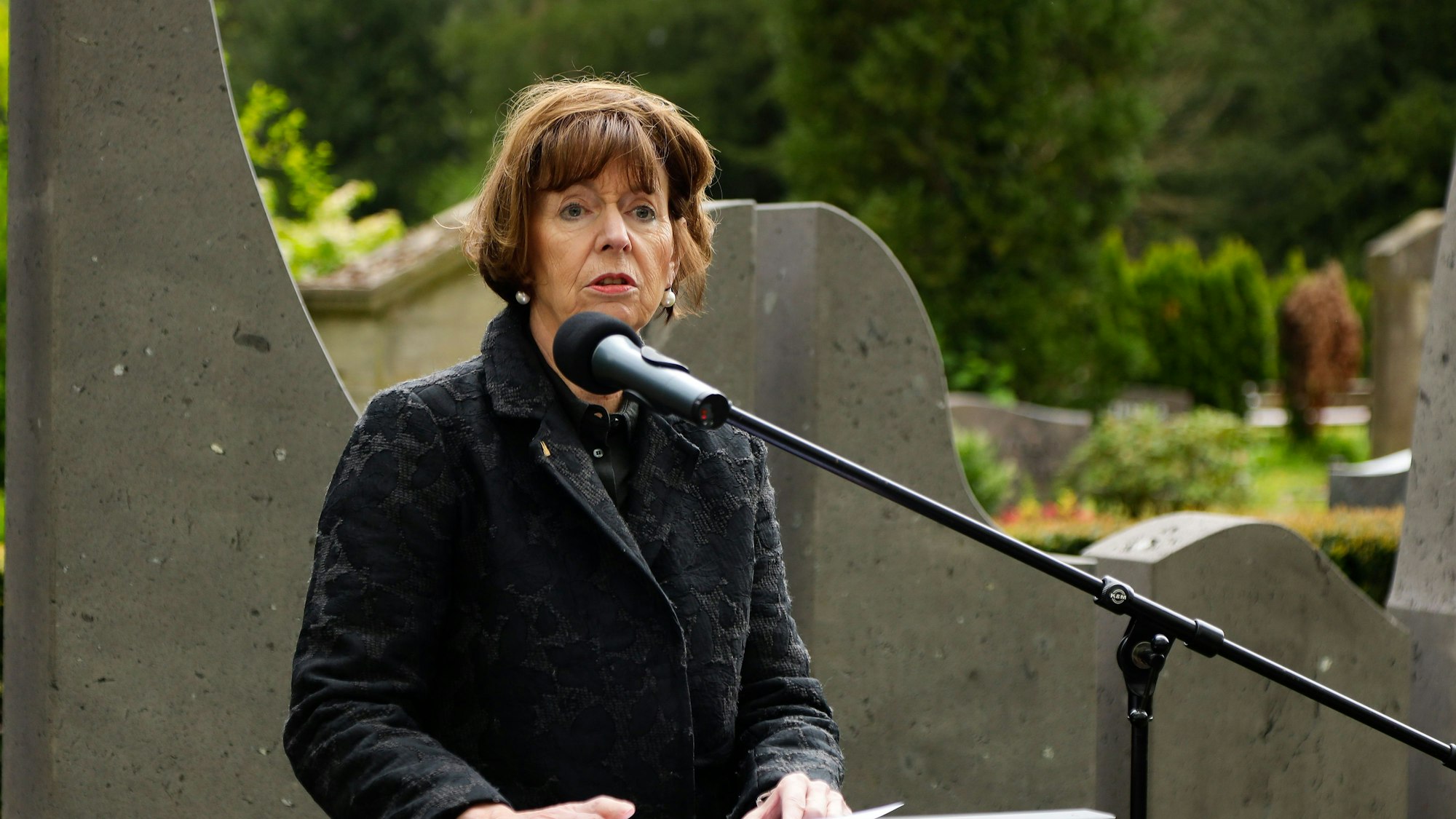 Henriette Reker an einem Rednerpult, sie spricht auf einem Friedhof mit ernster Miene in ein Mikrofon.