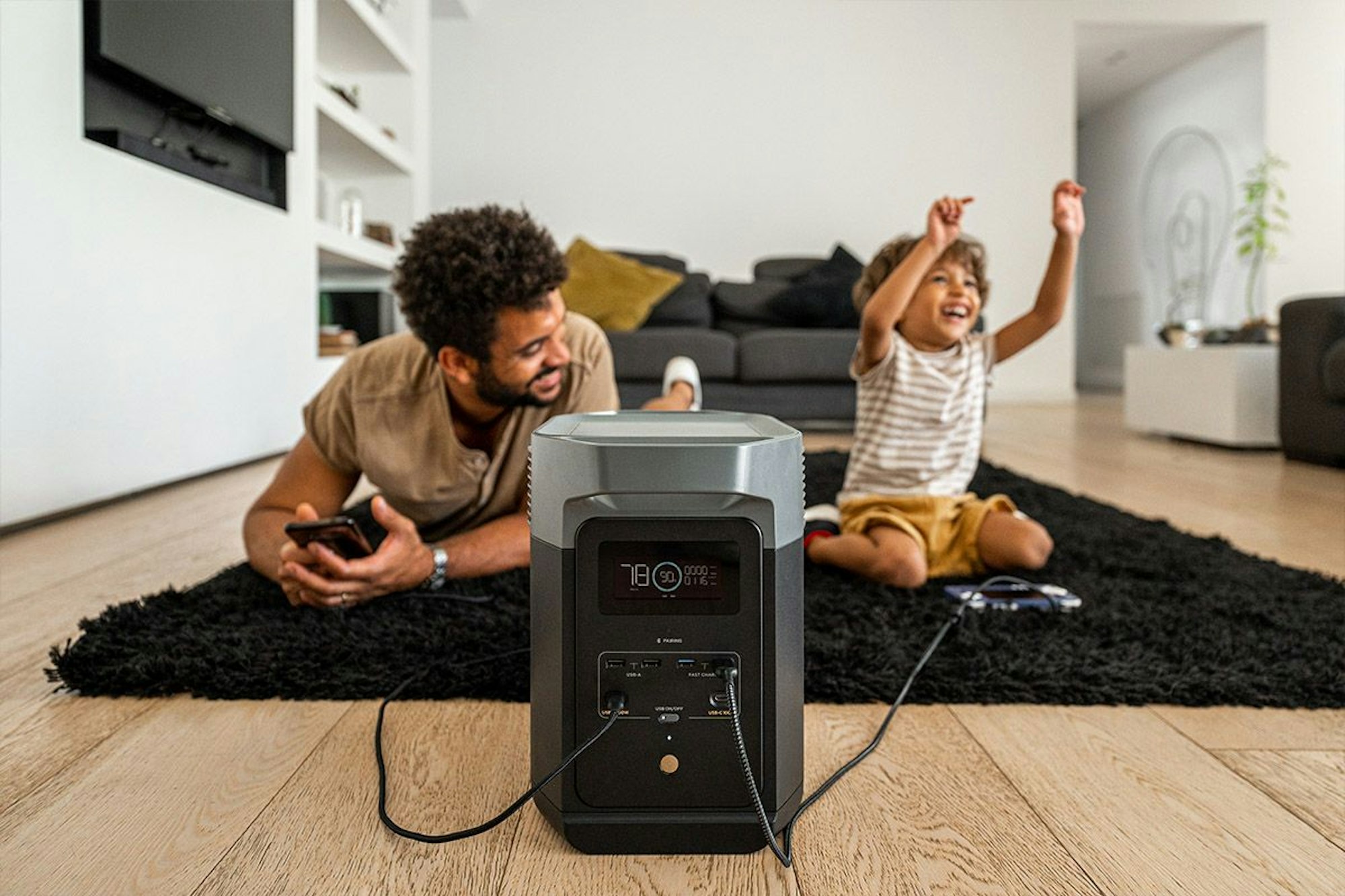 EcoFlow Powerstation mobiler Speicher steht auf dem Boden, im Hintergrund sitzen Vater und Sohn auf Teppichboden im Wohnzimmer.