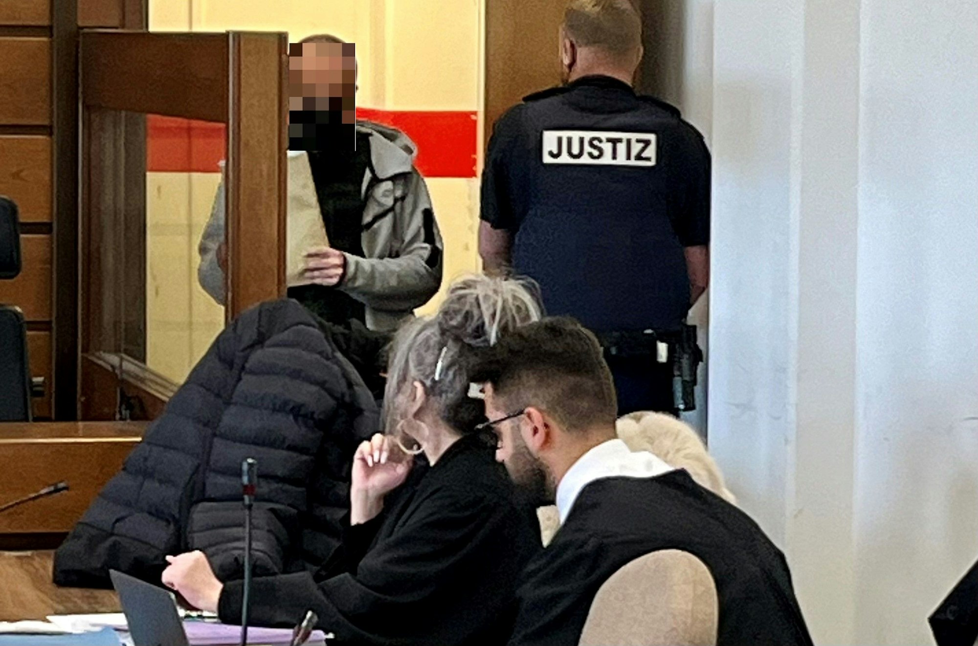 Ein Angeklagter betritt den Gerichtssaal, neben der Tür steht ein Justizwachtmeister. Ein weiterer Angeklagter sitzt neben seiner Anwältin und versteckt sich unter einer Jacke.
