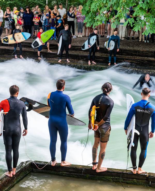 Dreizehn Surfer warten am Uferrand der Eisbachwelle in München