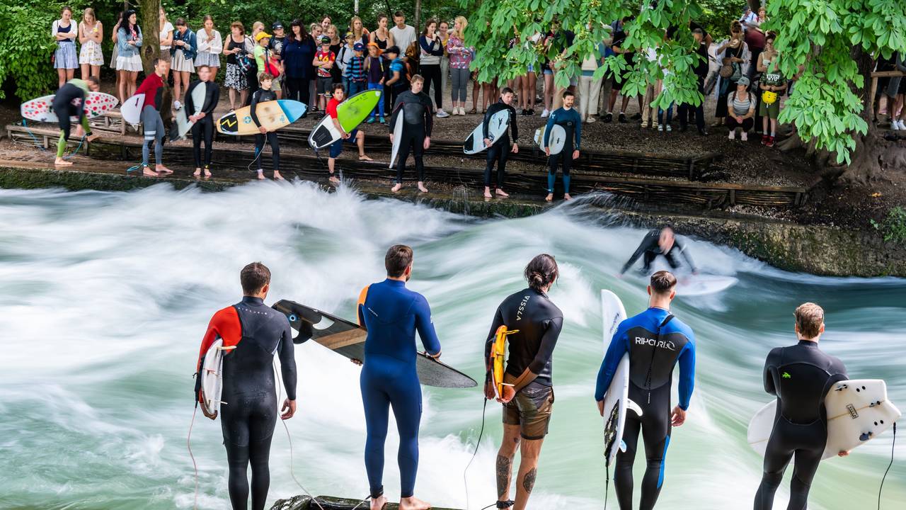 Dreizehn Surfer warten am Uferrand der Eisbachwelle in München