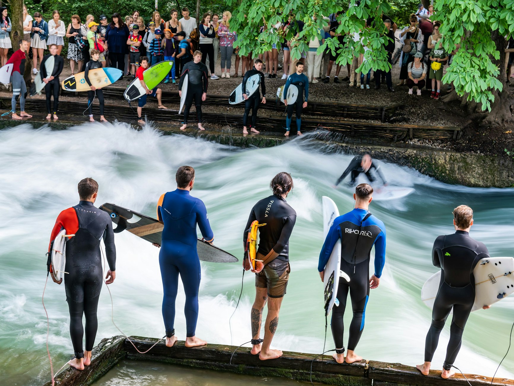 Dreizehn Surfer warten am Uferrand der Eisbachwelle in München