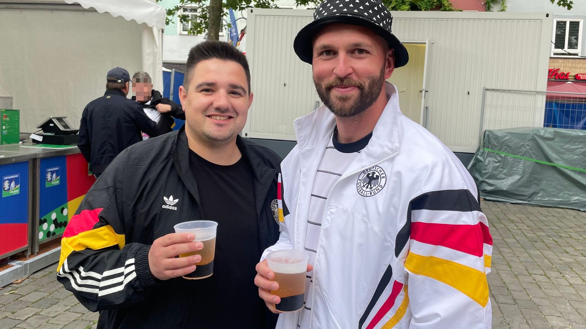 Zwei Männer halten ein Becher Bier in der Hand, beide tragen Jacken mit Streifen in den Deutschland-Farben.
