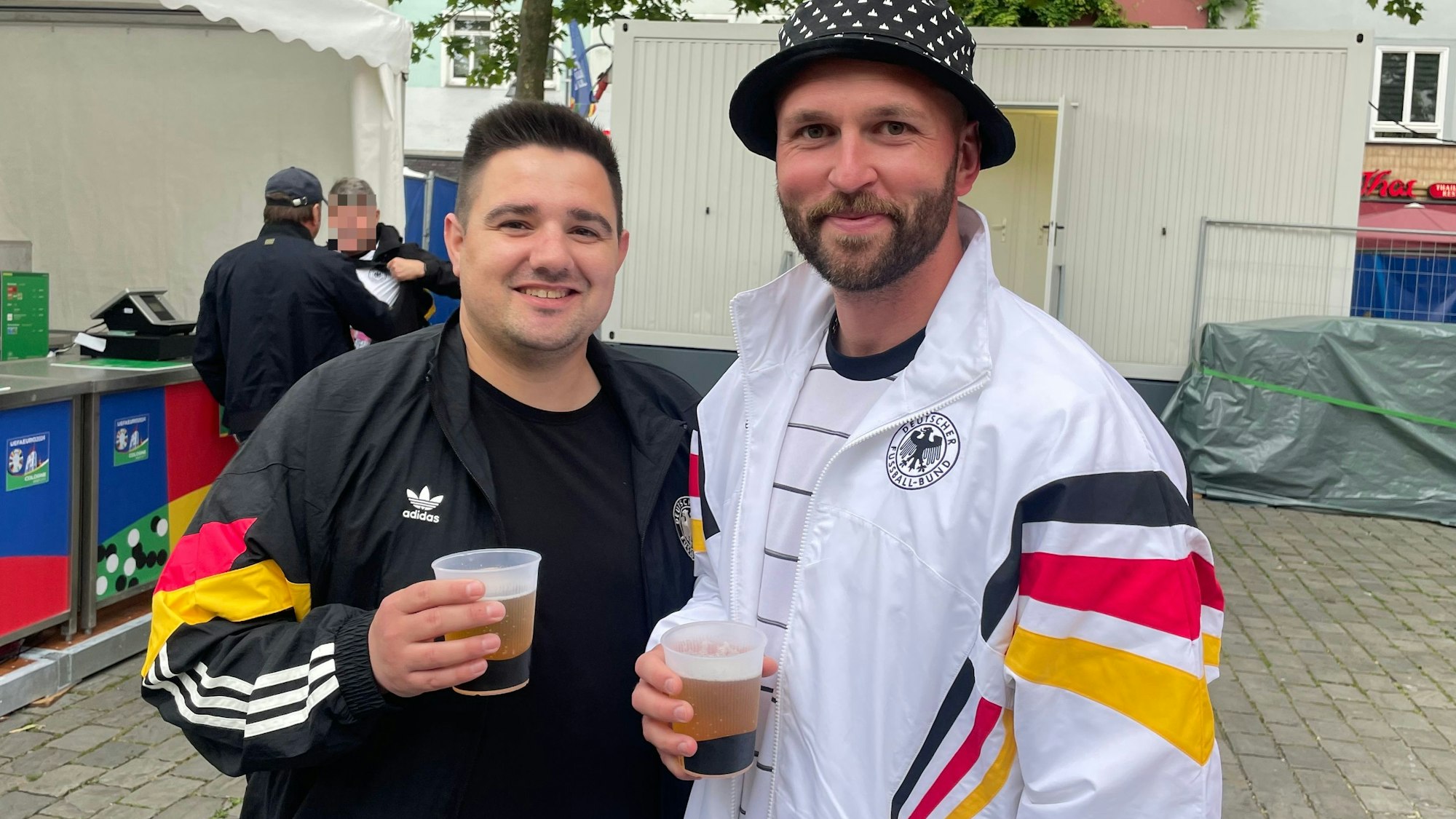Zwei Männer halten ein Becher Bier in der Hand, beide tragen Jacken mit Streifen in den Deutschland-Farben.