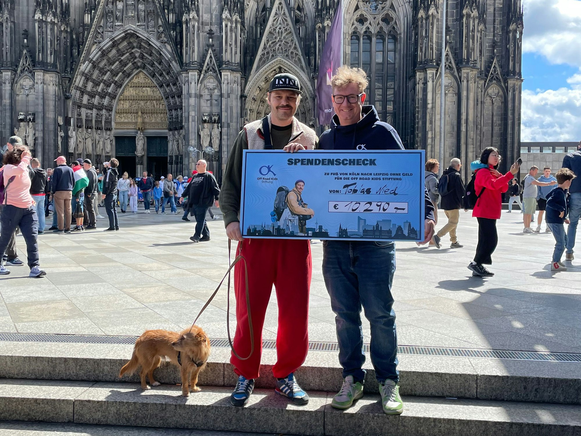 Tobias Nied und Tomi Czech stehen vor dem Kölner Dom mit einem Spendenscheck.