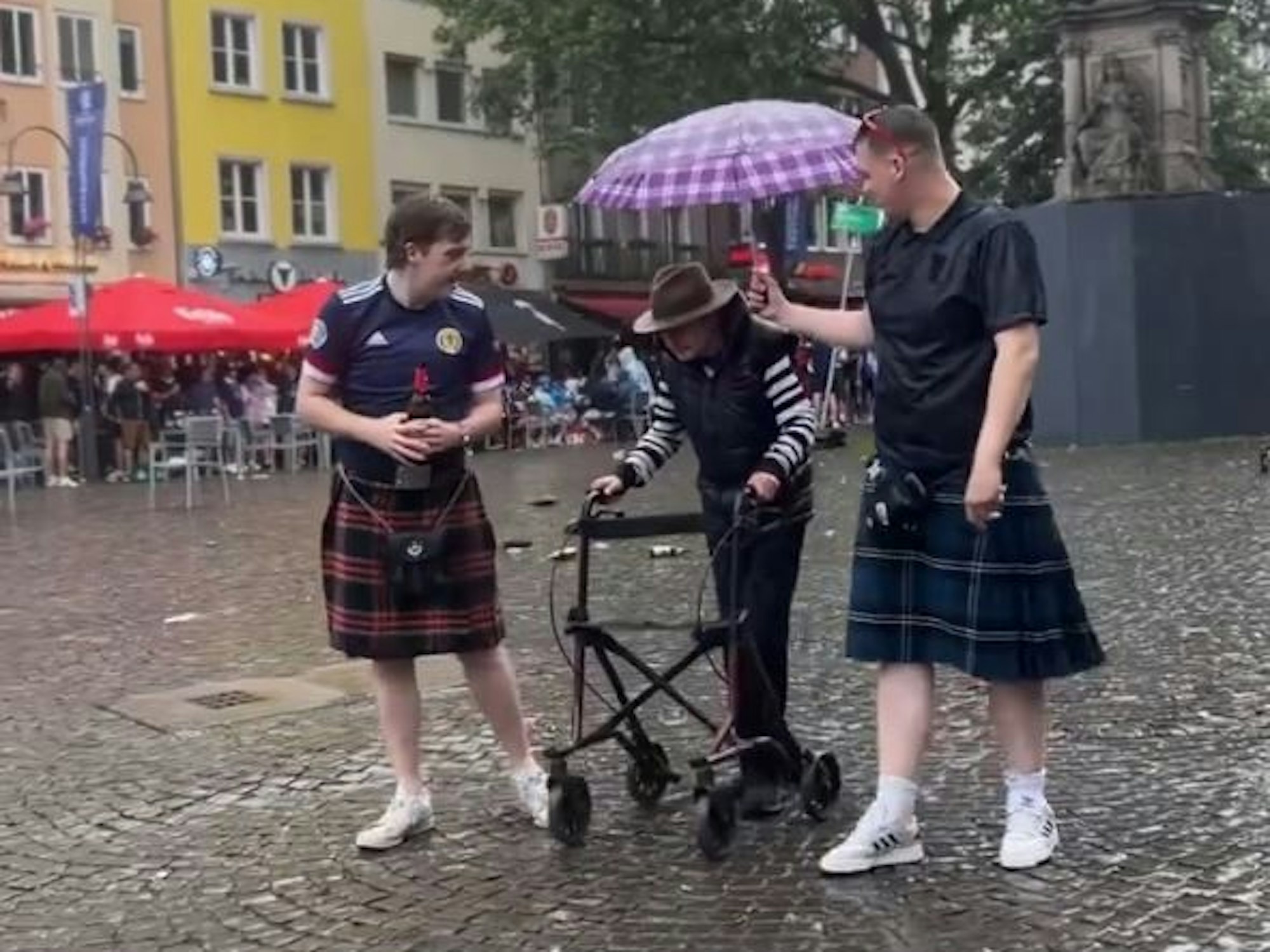 Zwei Männer mit Schottenrock begleiten einen Opa mit Gehhilfe und halten einen Regenschirm über ihn.