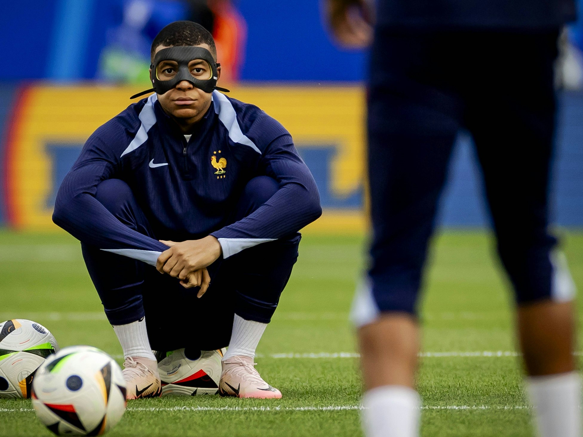 Kylian Mbappé sitzt mit Gesichtsmaske auf einem Fußball.