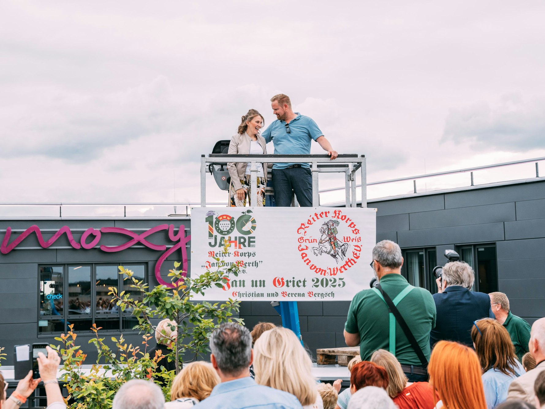 Ein Mann und eine Frau stehen auf einer Hebebühne vor anderen Menschen.