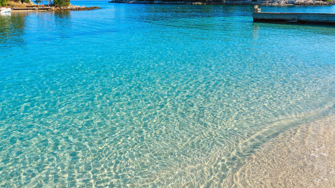 Wunderschönes Ionisches Meer mit klarem türkisfarbenem Wasser und morgendlichem Sommerblick auf die Küste vom Strand aus (Ksamil, Albanien).