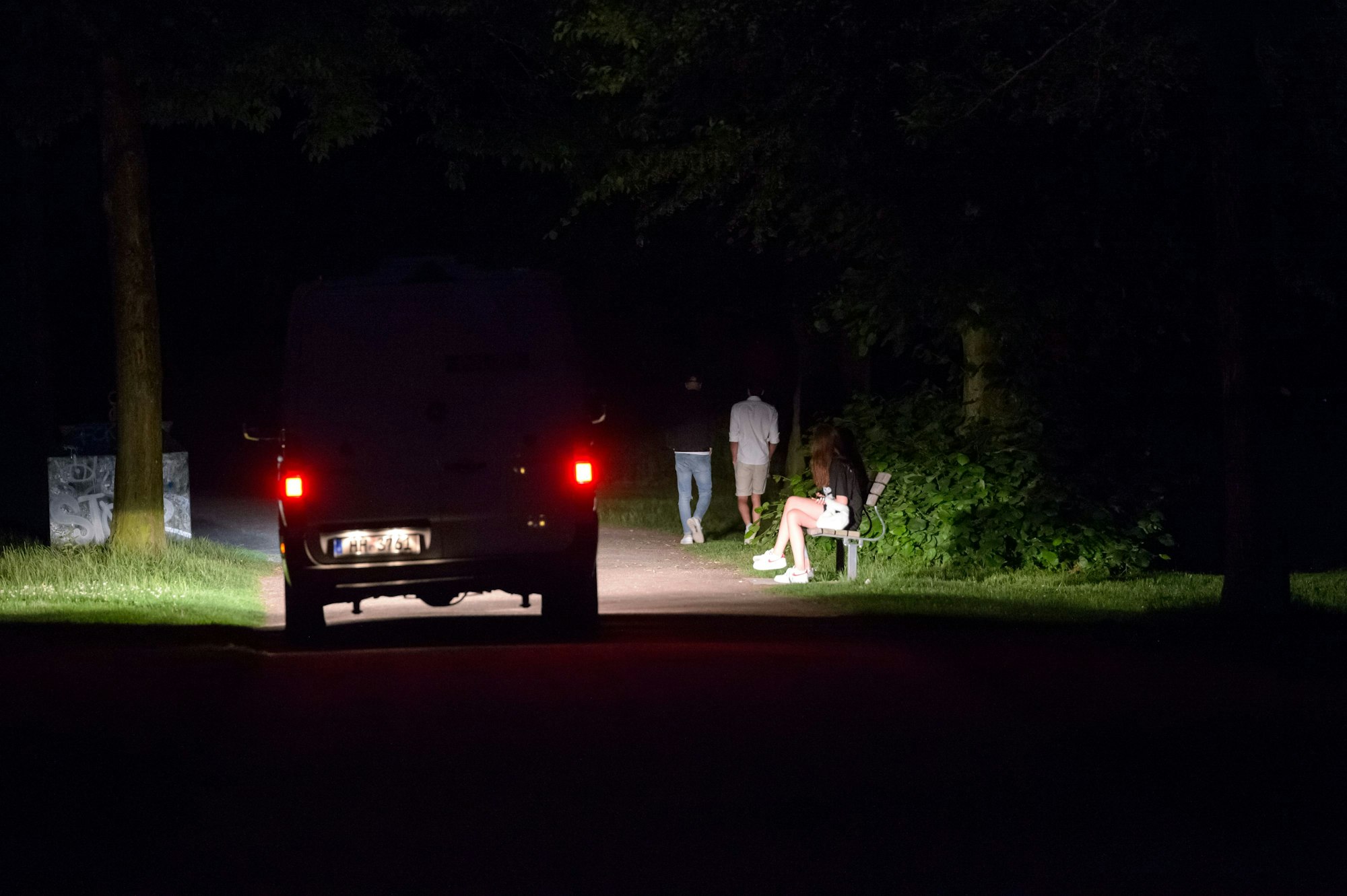 Ein Polizeifahrzeug fährt durch den dunklen, fast menschenleeren Stadtpark in Hamburg, während im Licht der Scheinwerfer vier Jugendliche stehen oder laufen.