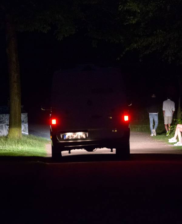 Ein Polizeifahrzeug fährt durch den dunklen, fast menschenleeren Stadtpark in Hamburg, während im Licht der Scheinwerfer vier Jugendliche stehen oder laufen.