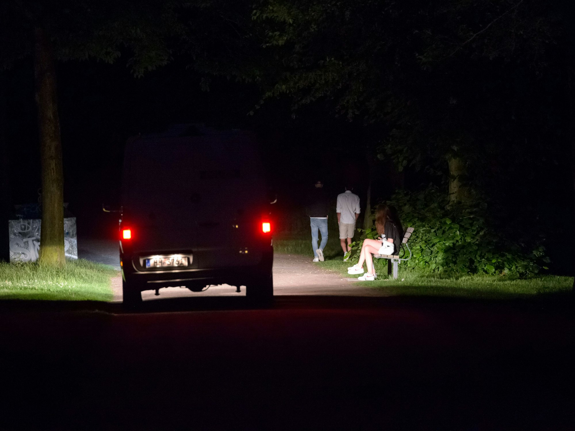 Ein Polizeifahrzeug fährt durch den dunklen, fast menschenleeren Stadtpark in Hamburg, während im Licht der Scheinwerfer vier Jugendliche stehen oder laufen.