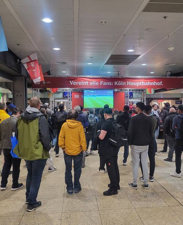 Einige Menschen stehen im Kölner Hauptbahnhof vor einem Fernseher und schauen gemeinsam ein EM-Spiel.