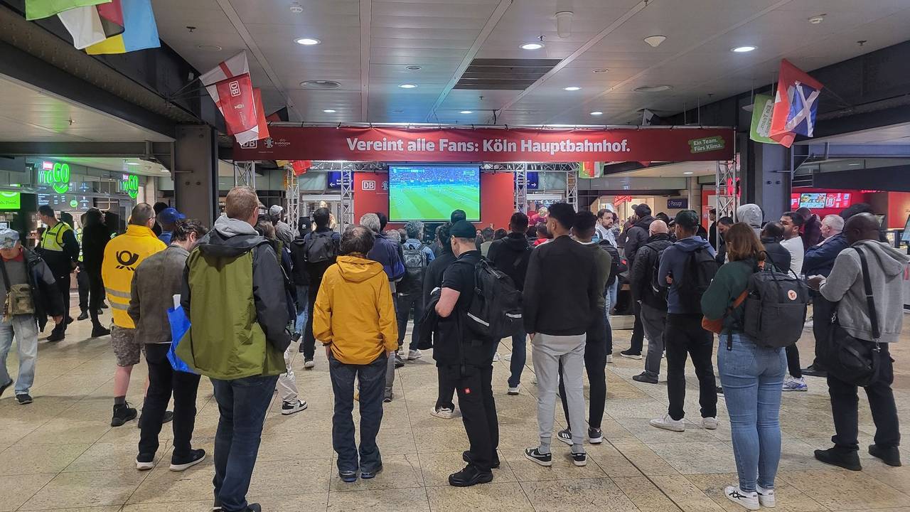 Einige Menschen stehen im Kölner Hauptbahnhof vor einem Fernseher und schauen gemeinsam ein EM-Spiel.