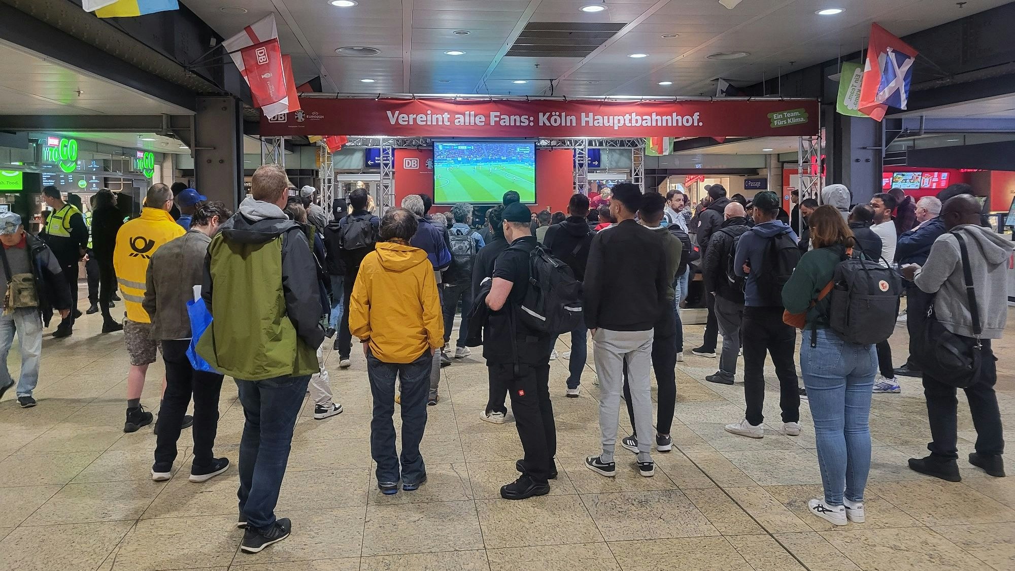 Einige Menschen stehen im Kölner Hauptbahnhof vor einem Fernseher und schauen gemeinsam ein EM-Spiel.