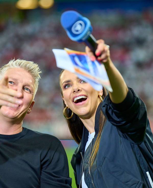 Bastian Schweinsteiger und Esther Sedlaczek blicken am Rande einer Übertragung zur EM 2024 lachend Richtung Tribüne.
