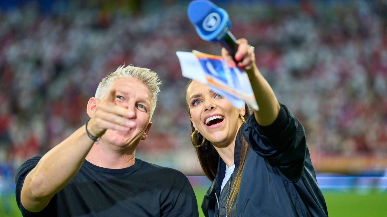 Bastian Schweinsteiger und Esther Sedlaczek blicken am Rande einer Übertragung zur EM 2024 lachend Richtung Tribüne.