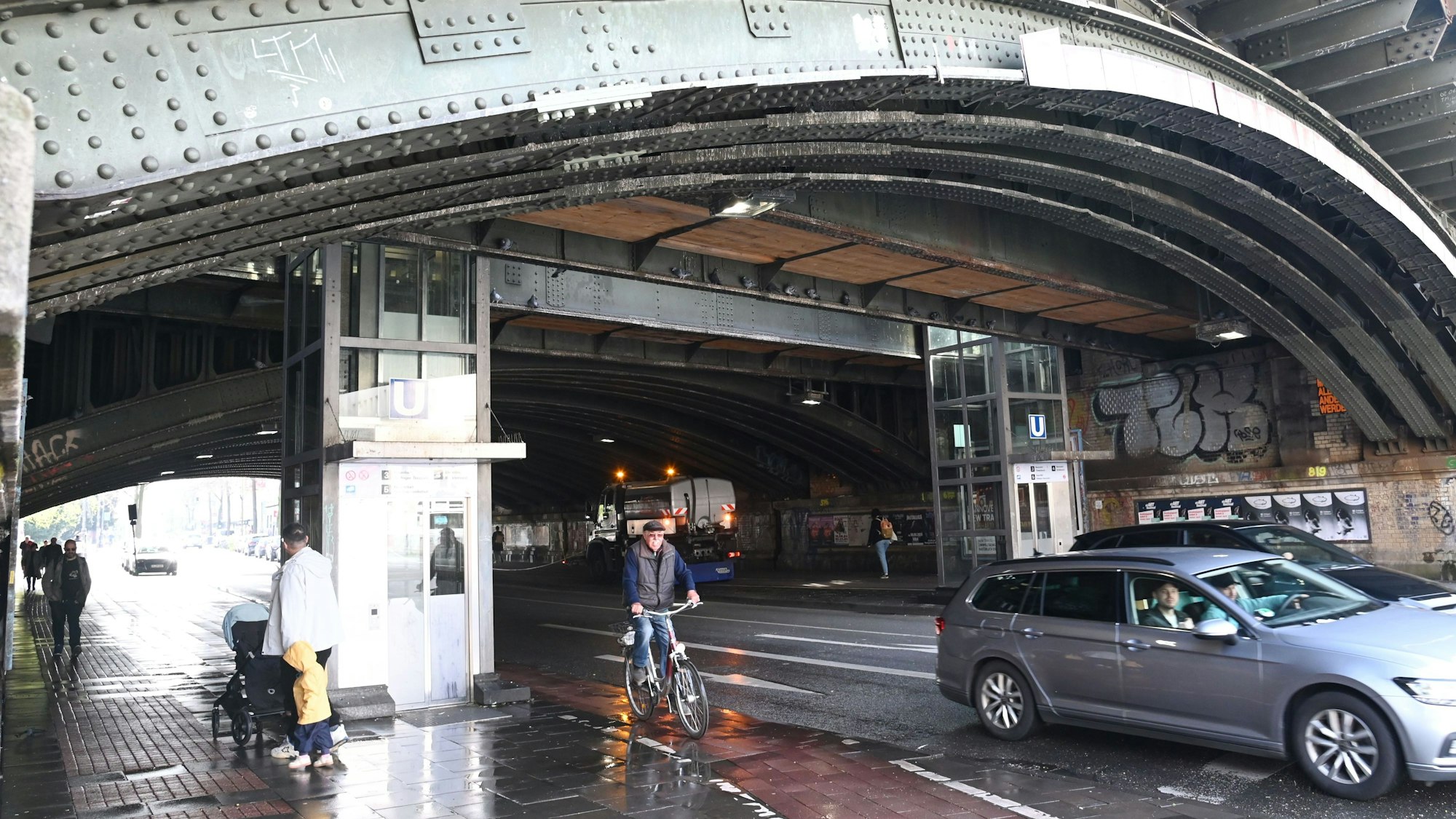 Autos und Fahrräder fahren unter einem Bahnübergang entlang, auch einige Fußgänger sind zu sehen.