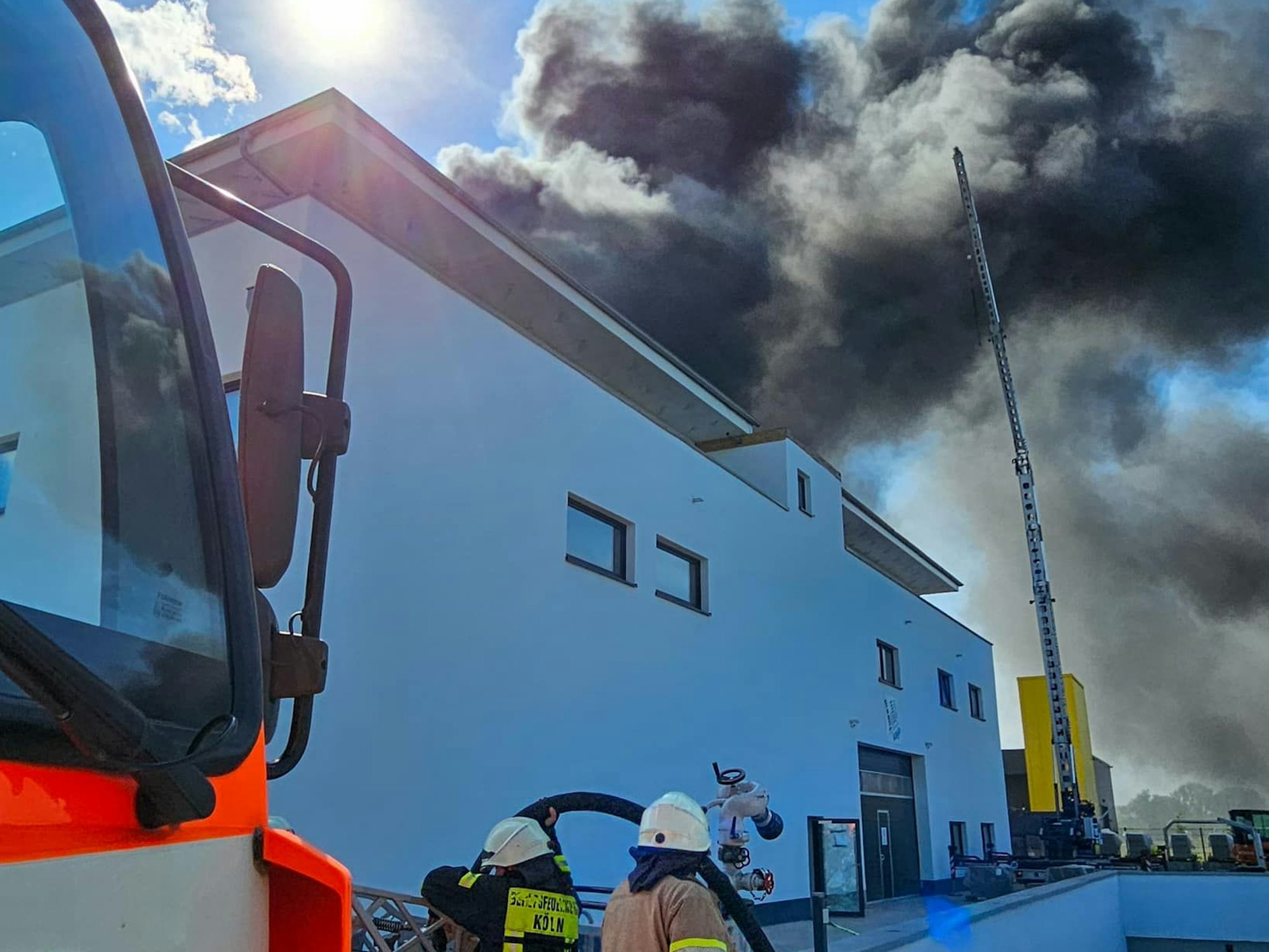 Vom Dach eines Gebäudes steigt dunkler Qualm auf. Vor dem Gebäude stehen ein Fahrzeug der Feuerwehr sowie zwei Einsatzkräfte.