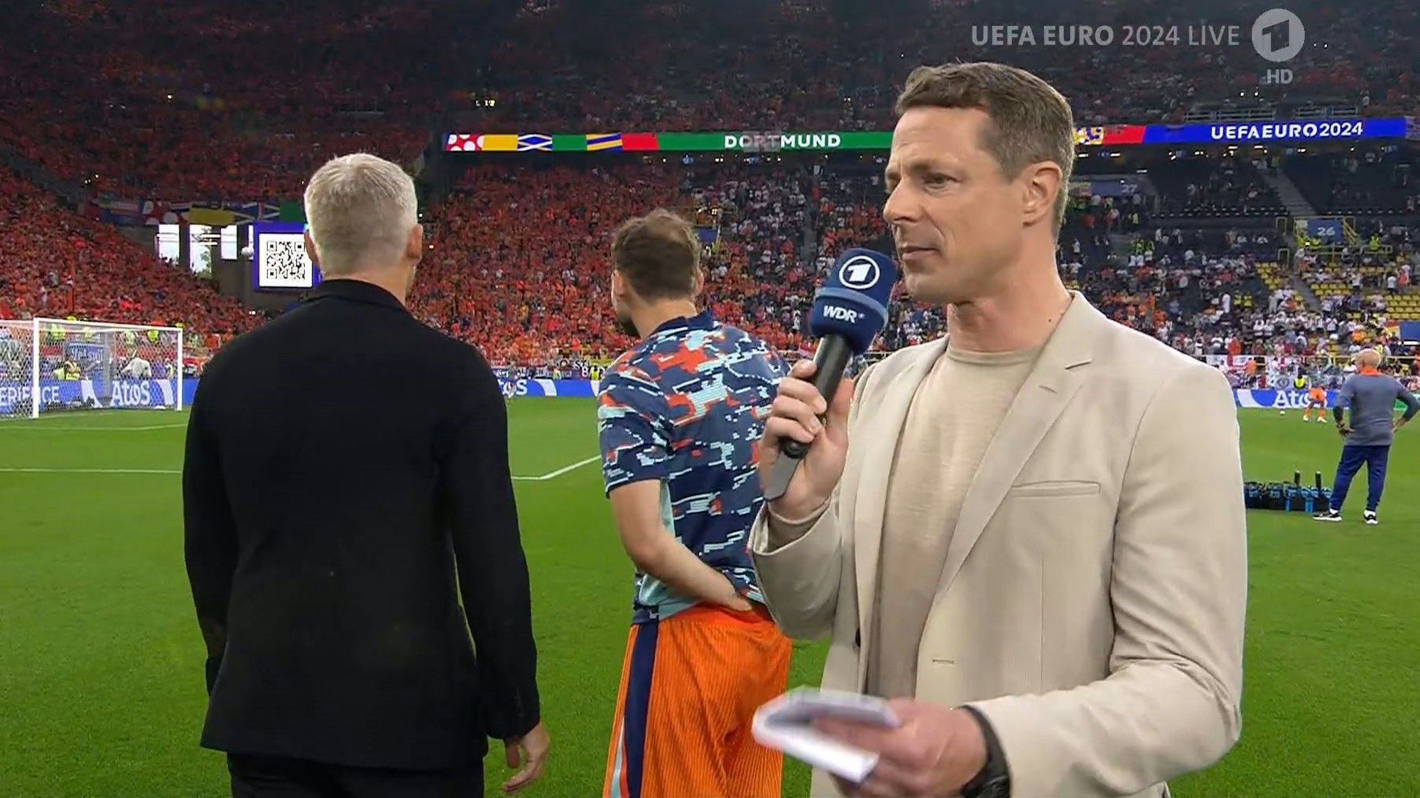 Alexander Bommes am 10. Juli 2024 beim EM-Halbfinale in der ARD-Übertragung. Im Hintergrund dreht ihm Bastian Schweinsteiger den Rücken zu.