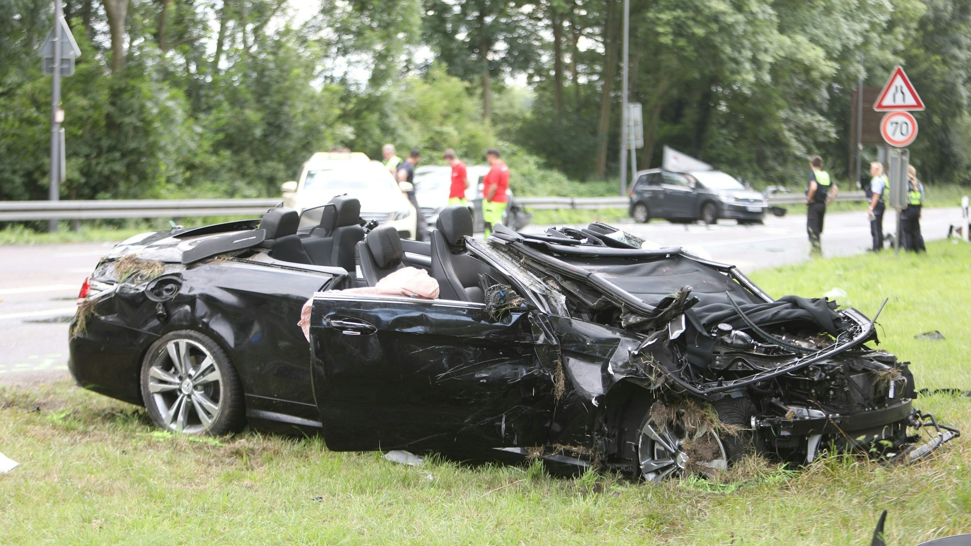 Ein stark beschädigtes Auto steht neben der Fahrbahn auf einem Grünsteifen. Das Dach fehlt.