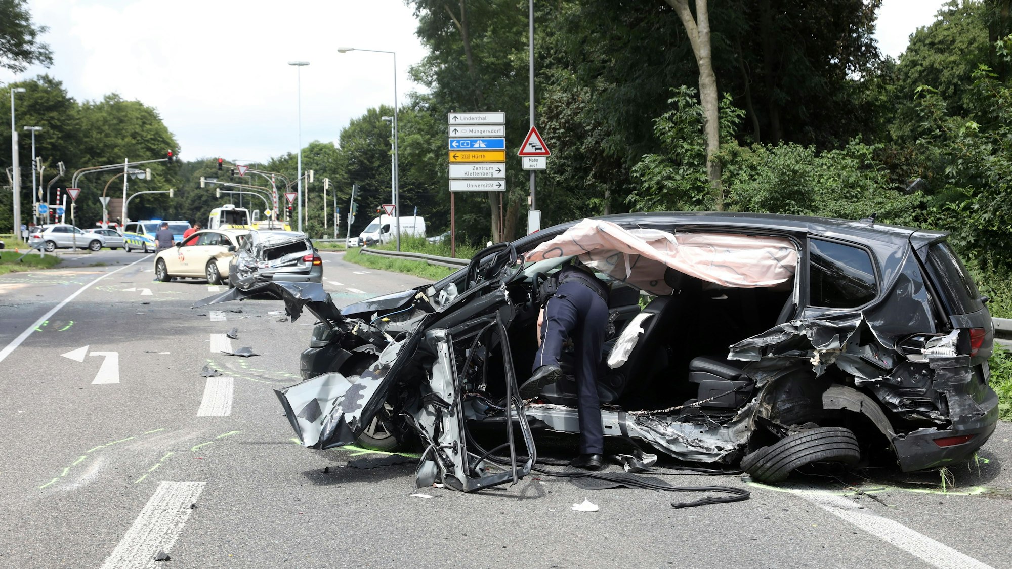Ein Beamter beugt sich in ein völlig zerfetztes Auto, im Hintergrund steht ein weiteres stark beschädigtes Fahrzeug auf der Fahrbahn.