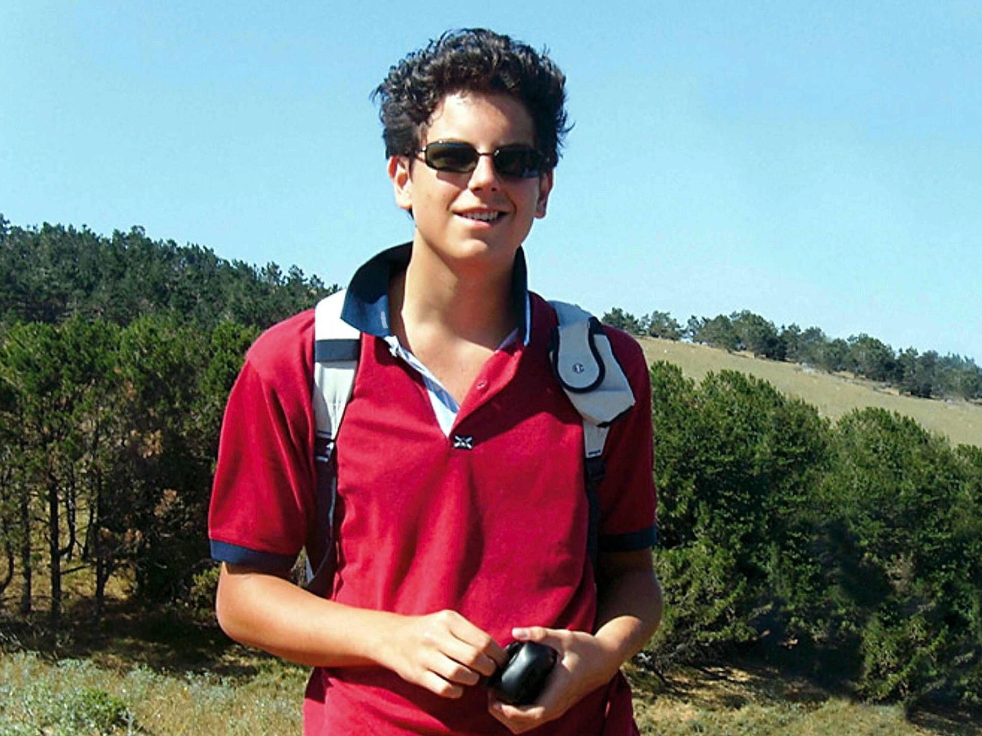 Carlo Acutis steht in einem roten Poloshirt und mit Sonnenbrille vor einem Wald.