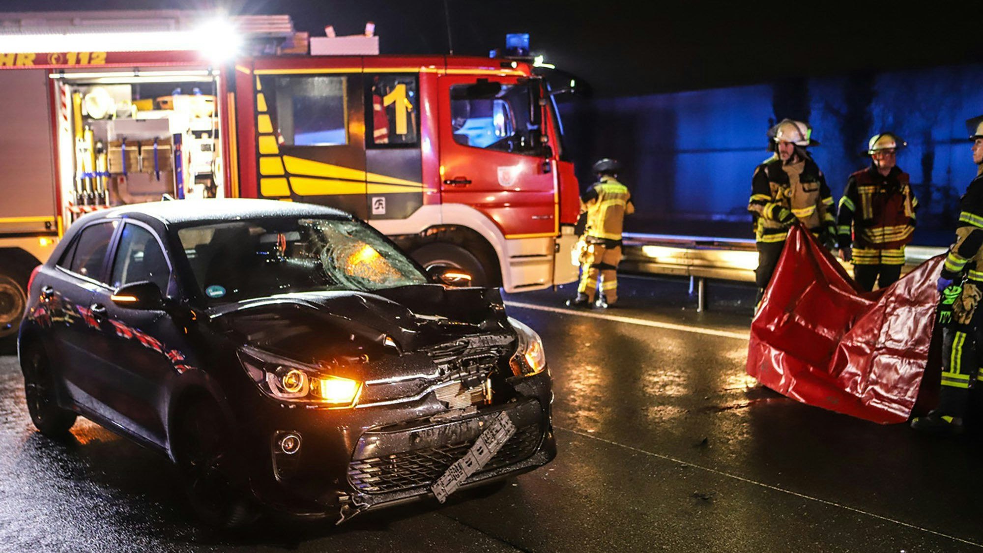 Einsatzkräfte der Feuerwehr stehen neben einem stark beschädigten Auto.