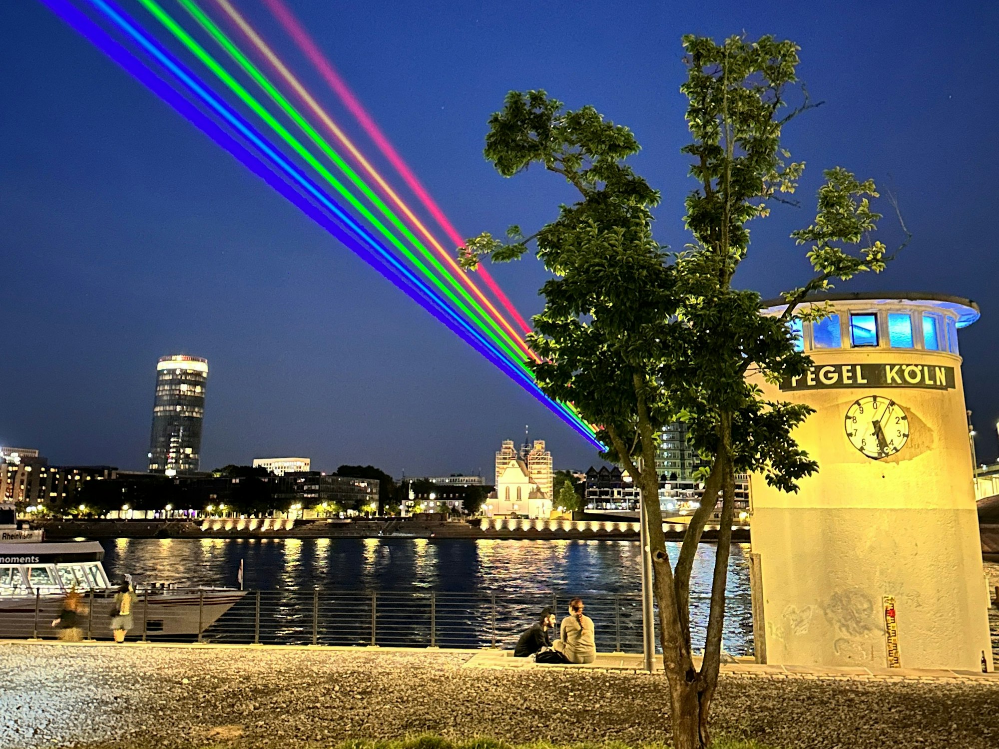 Laser-Regenbogen strahlt über Köln, im Vordergrund ist der Pegel Köln zu sehen.