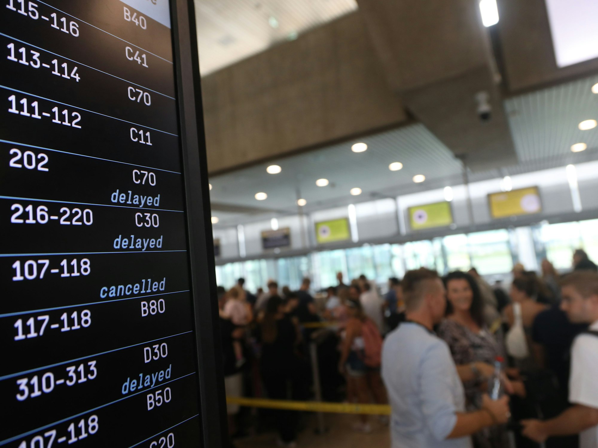 Eine Anzeigentafel am Flughafen zeigt verspätete und gestrichene Flüge an, im Hintergrund stehen zahlreiche Menschen.