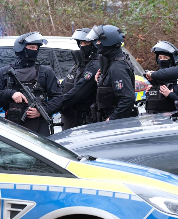 Schwer bewaffnete und vermummte Einsatzkräfte stehen hinter einem Streifenwagen.