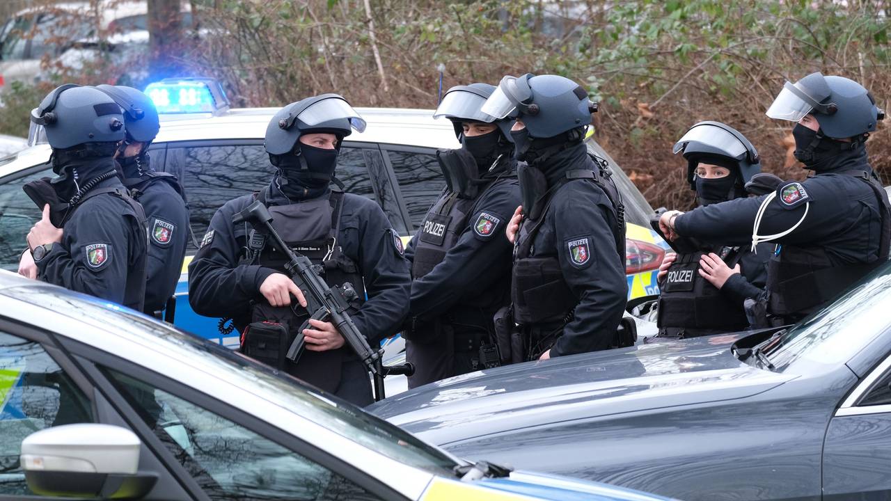 Schwer bewaffnete und vermummte Einsatzkräfte stehen hinter einem Streifenwagen.