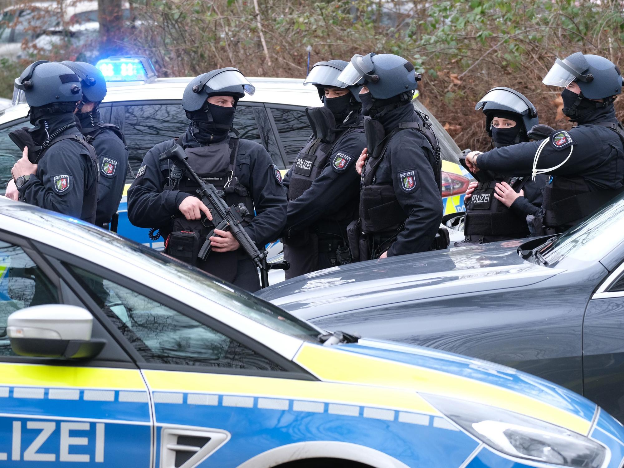 Schwer bewaffnete und vermummte Einsatzkräfte stehen hinter einem Streifenwagen.