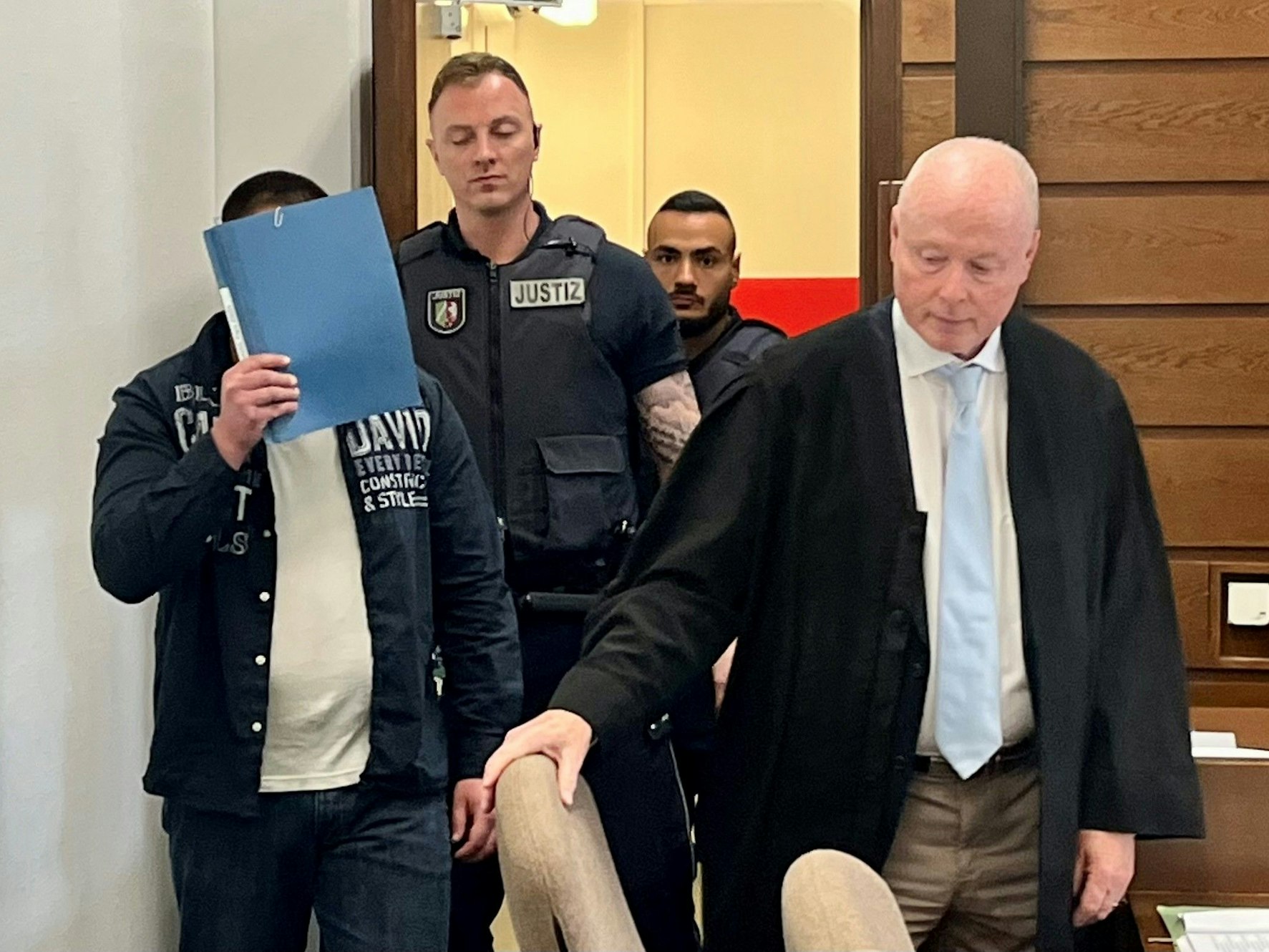 Ein Mann, der sein Gesicht hinter einer Mappe versteckt, wird von zwei Justizwachtmeistern in den Gerichtssaal gebracht. Sein Anwalt rückt ihm den Stuhl zurecht.