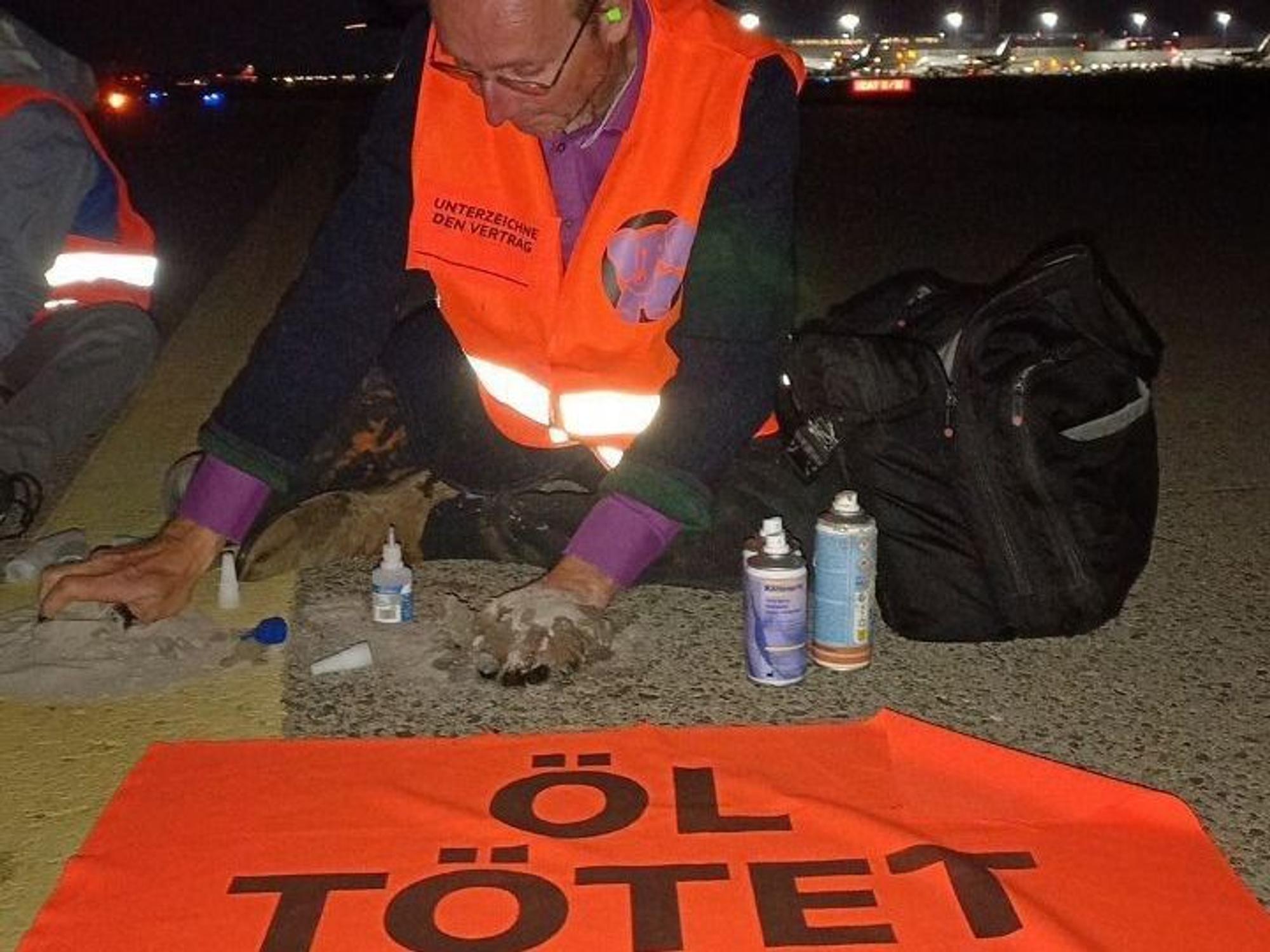 Ein Mann hat sich mit einer Hand auf dem Rollfeld festgeklebt, vor ihm ein Banner mit der Aufschrift „Öl tötet“.