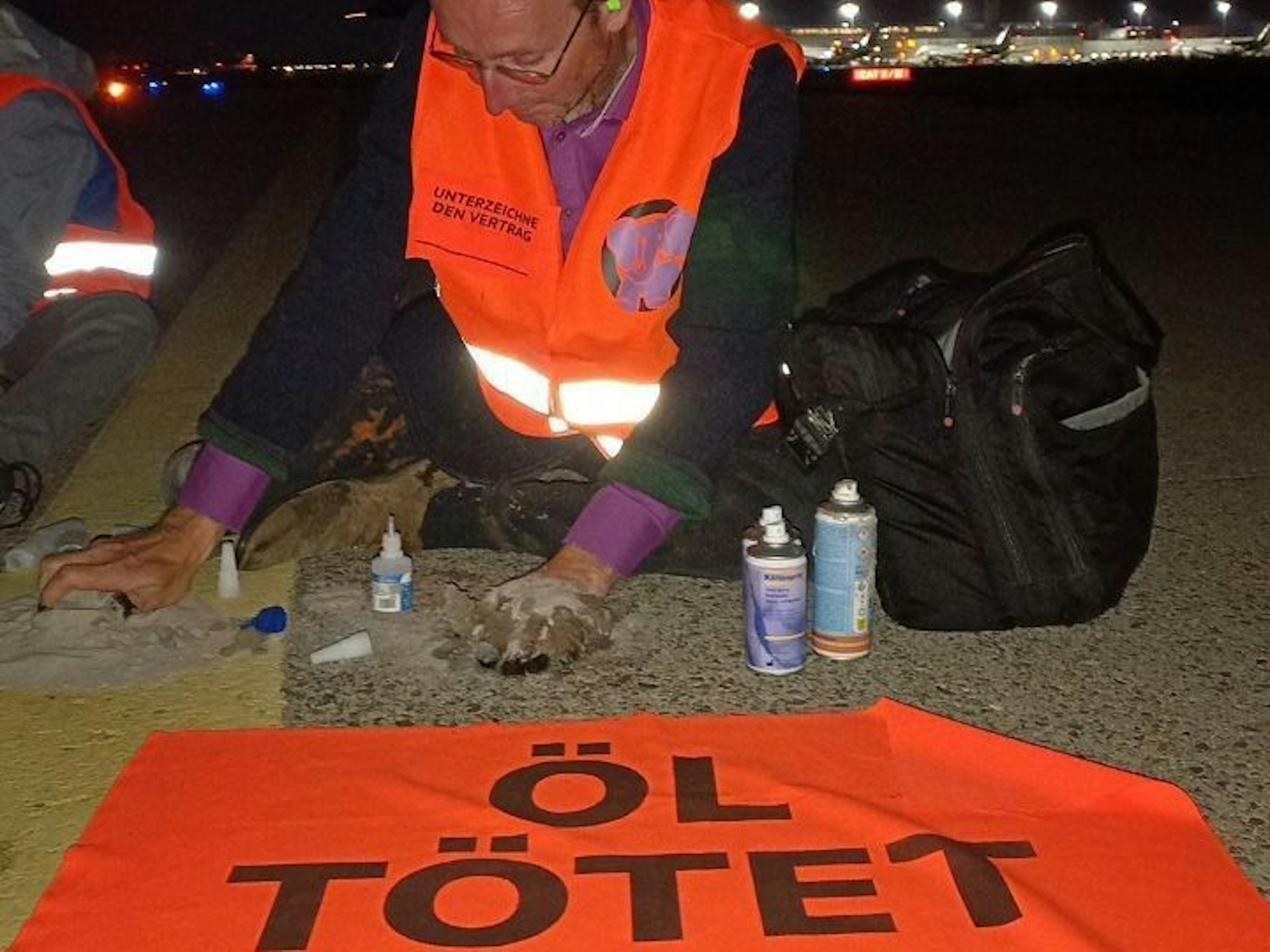 Ein Mann hat sich mit einer Hand auf dem Rollfeld eines Flughafens festgeklebt, vor ihm ein Banner mit der Aufschrift „Öl tötet“.