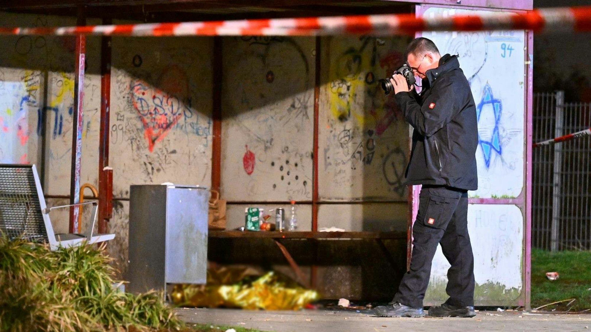 Ein Leichnam liegt abgedeckt in einem Wartehäuschen, ein Polizist macht Fotos.