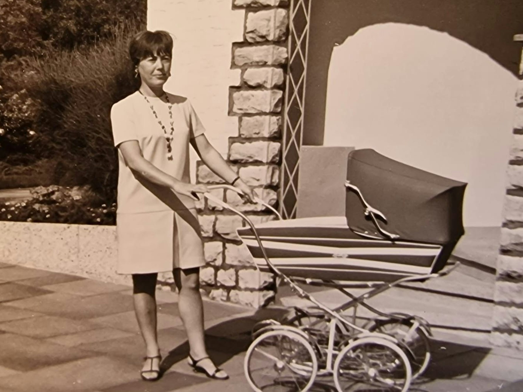 Auf einem alten Schwarz-Weiß-Foto steht eine junge Frau vor einem Kinderwagen.