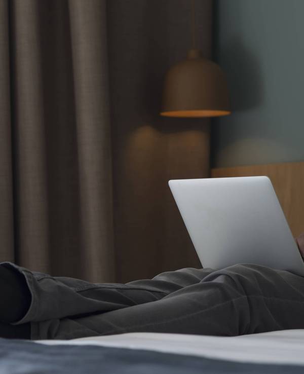 Ein Mann sitzt mit seinem Laptop auf einem Hotelbett.