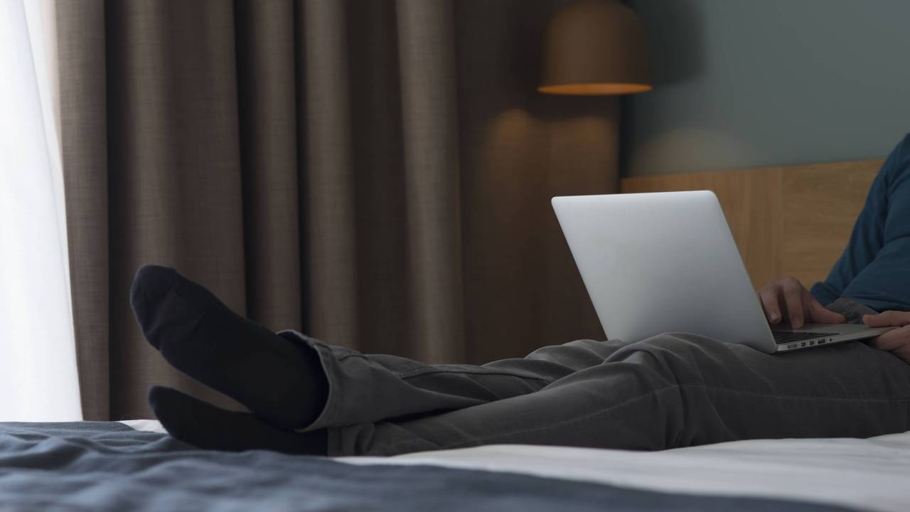 Ein Mann sitzt mit seinem Laptop auf einem Hotelbett.