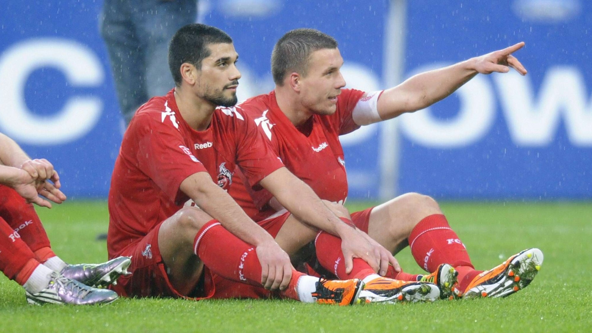 Taner Yalcin und Lukas Podolski sitzen zusammen auf dem Rasen des Rhein-Energie-Stadions.