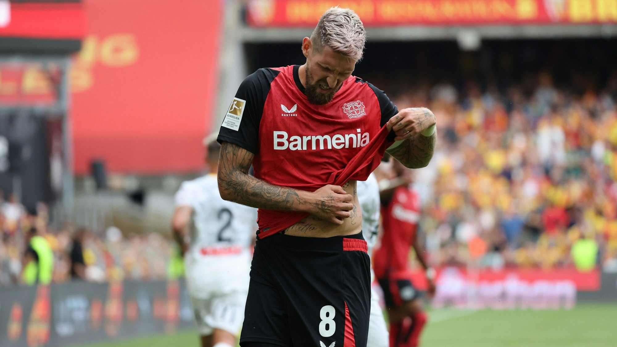 Robert Andrich kratzt sich bei einem Spiel von Bayer Leverkusen den Bauch. Er trägt das neue Trikot der Werkself.