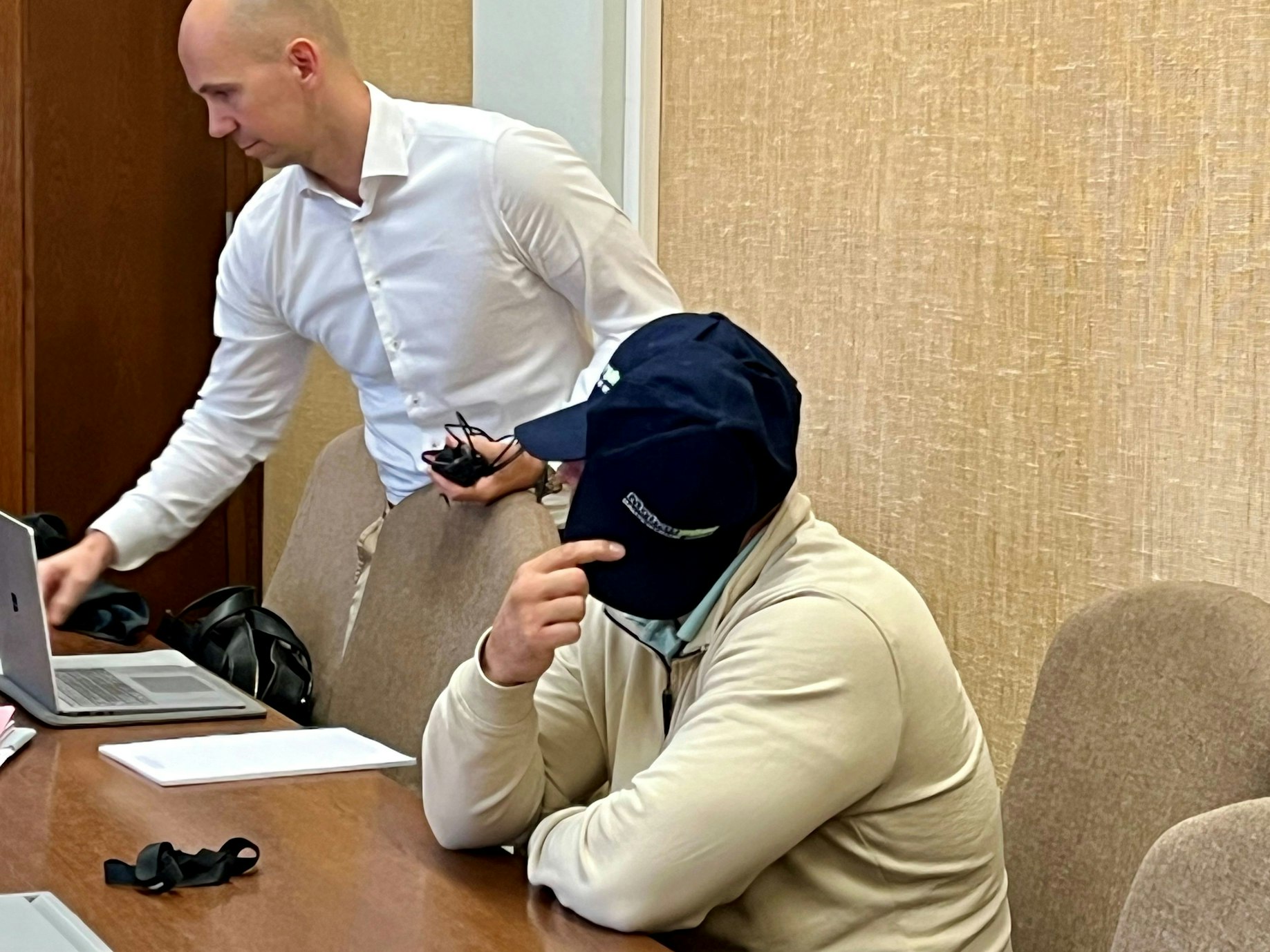 Ein Mann sitzt an einem Tisch und verdeckt sein Gesicht mit einer Baseballkappe. Neben ihm steht ein anderer Mann und beugt sich zu seinem Laptop.