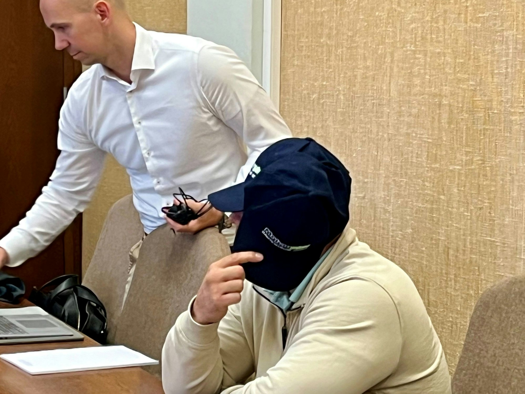 Ein Mann sitzt auf der Anklagebank, er trägt eine Basecap und hält sich eine zweite vor das Gesicht. Neben ihm steht sein Anwalt.