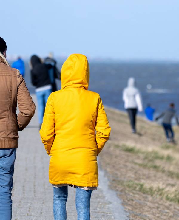 Touristen und Touristinnen gehen bei sonnigem Wetter auf einem Deich am Hafen entlang
