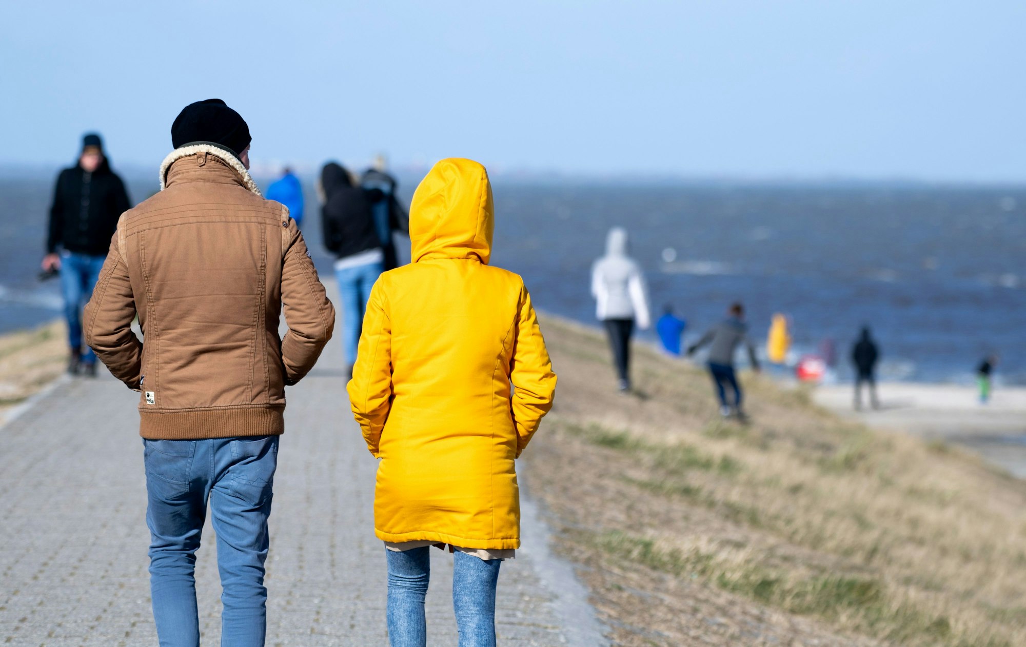 Touristen und Touristinnen gehen bei sonnigem Wetter auf einem Deich am Hafen entlang