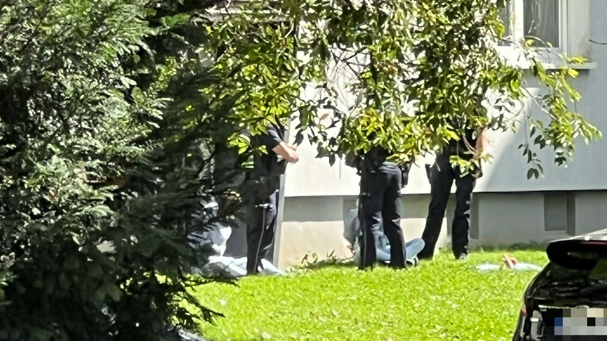 Zwei Personen sitzen mit auf dem Rücken gefesselten Händen vor einem Haus auf dem Rasen, vor ihnen stehen drei Polizisten.