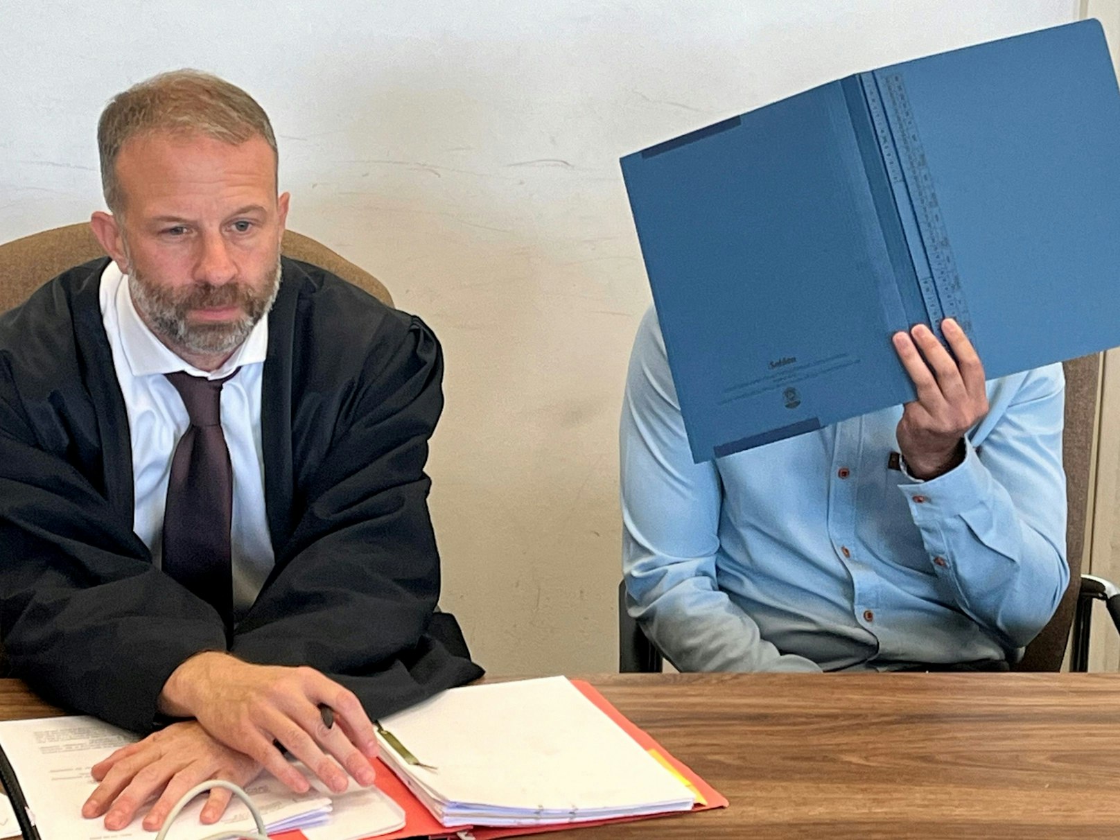 Ein Mann hält sich eine Mappe vor das Gesicht, neben ihm sitzt sein Anwalt.