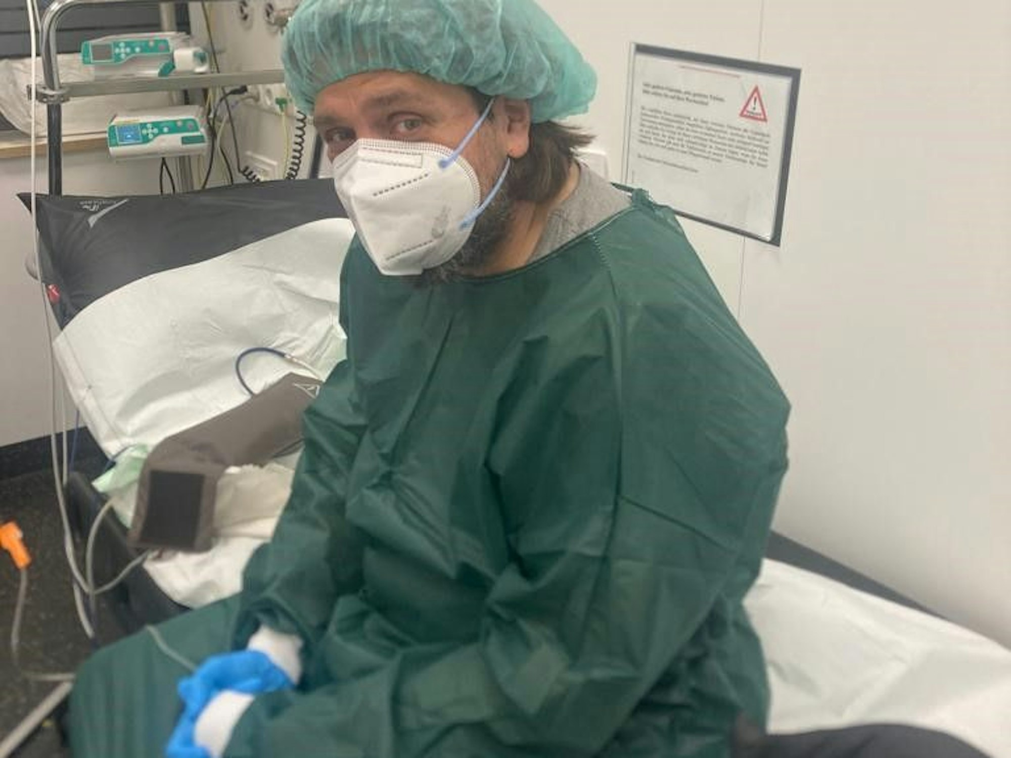 Thomas Haensgen sitzt in Schutzkleidung und mit Mundschutz im Krankenhaus auf einer Liege.