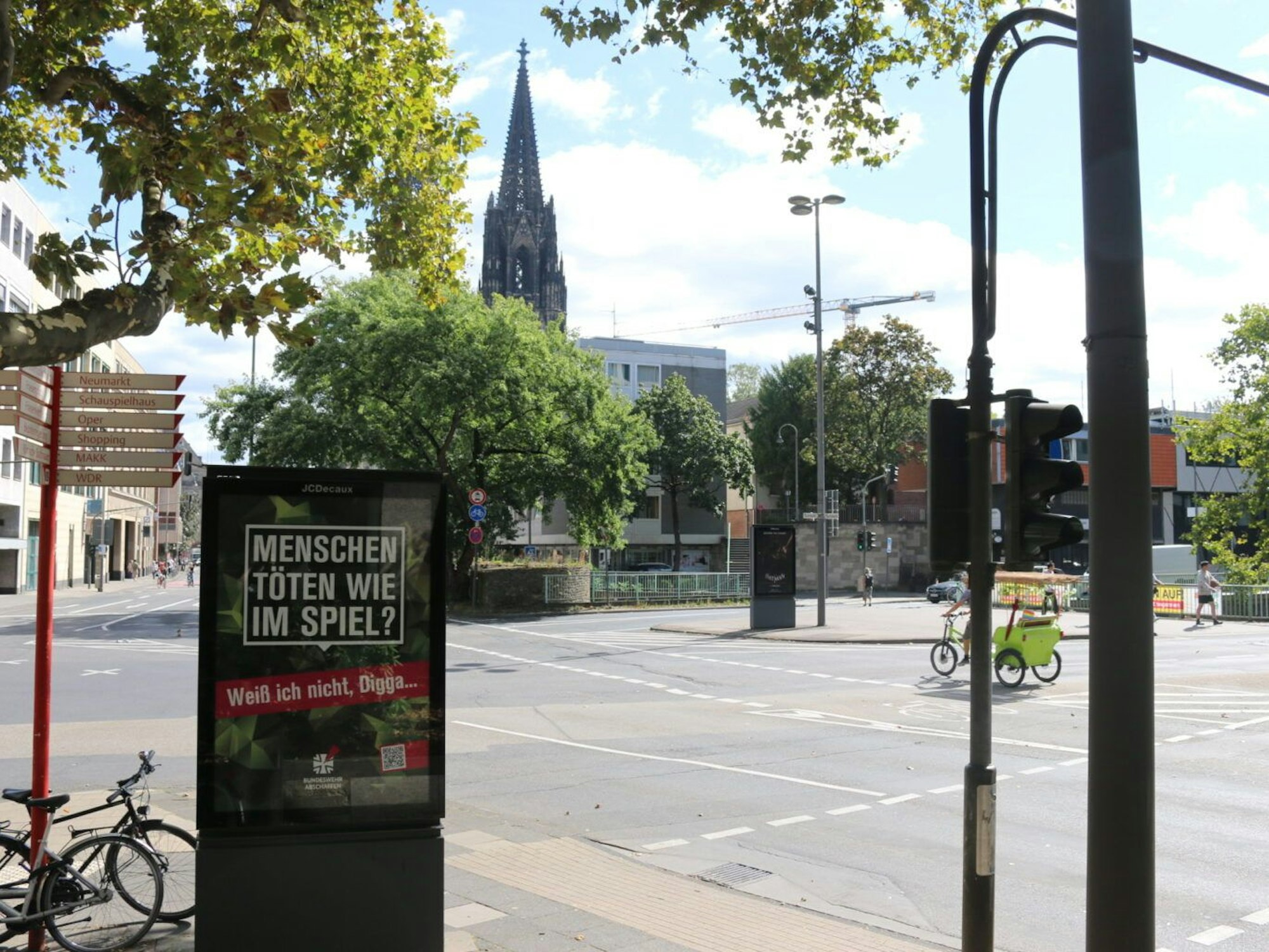 Ein Plakat mitten im Straßenverkehr von Köln, im Hintergrund ist der Kölner Dom zu sehen.